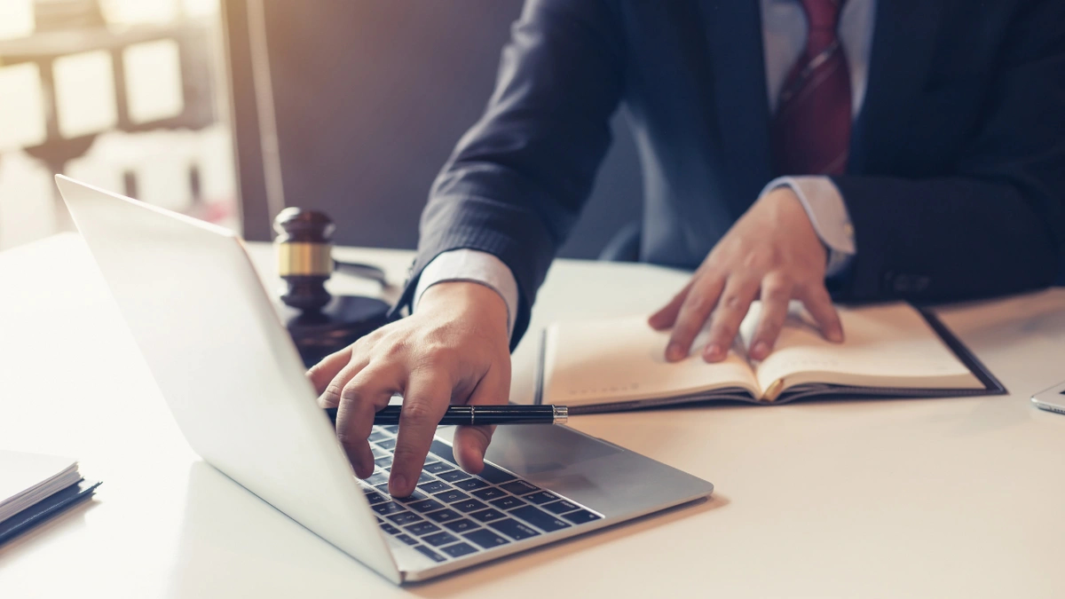 Lawyer wearing a suit looking at a ledger and typing on a laptop with a gavel on the desk.