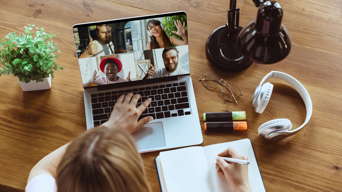 Birds eye view of a person sitting at a wooden desk writing in a journal while typing on a laptop where four colleagues are on a video call.