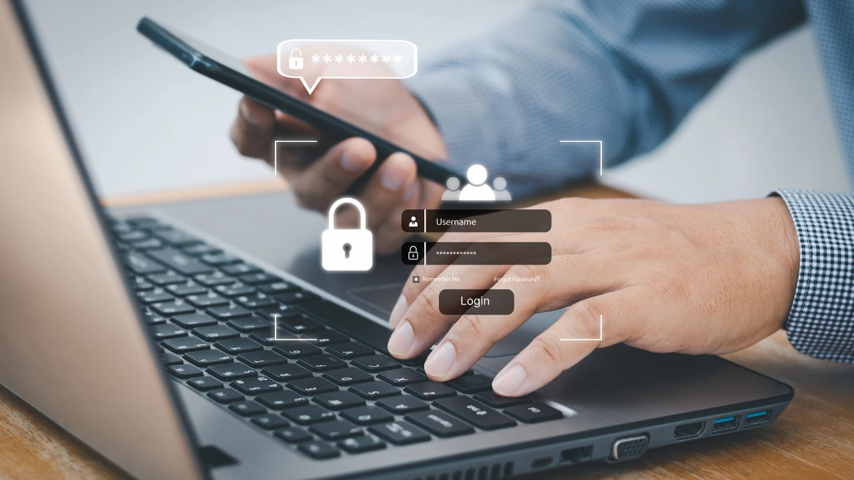 A man typgin on a laptop while holding his phone, there are different security icons overlayed including a log in screen and a lock and masked password.