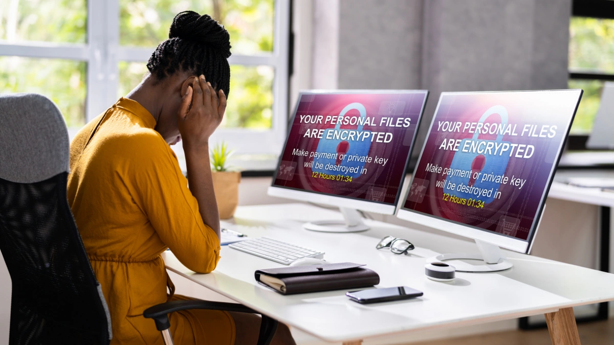 A woman sitting at a desk holds her head in her hands while looking at two computer screens that both say "Your Personal Files Are Encrypted"