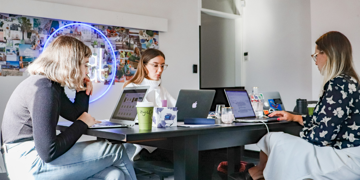 Three young women sitting around a table on macbooks. Each doing something different.