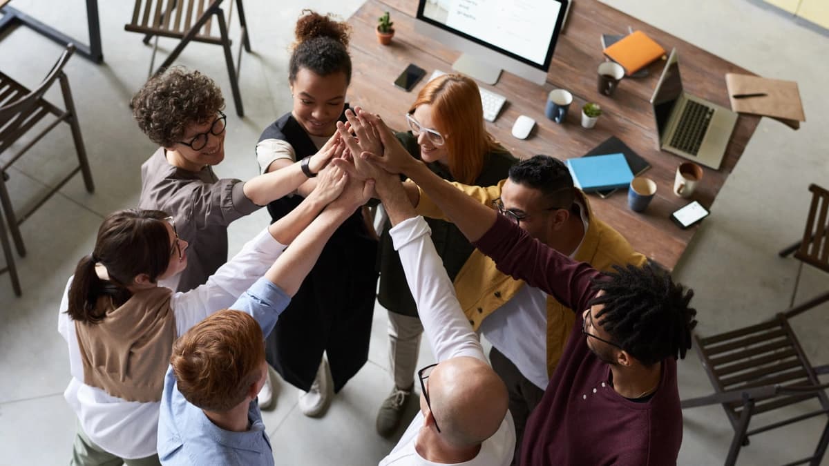 A team of diverse workers standing in a circle with their arms outstretched and hands touching, there is a desk with computers in the background.