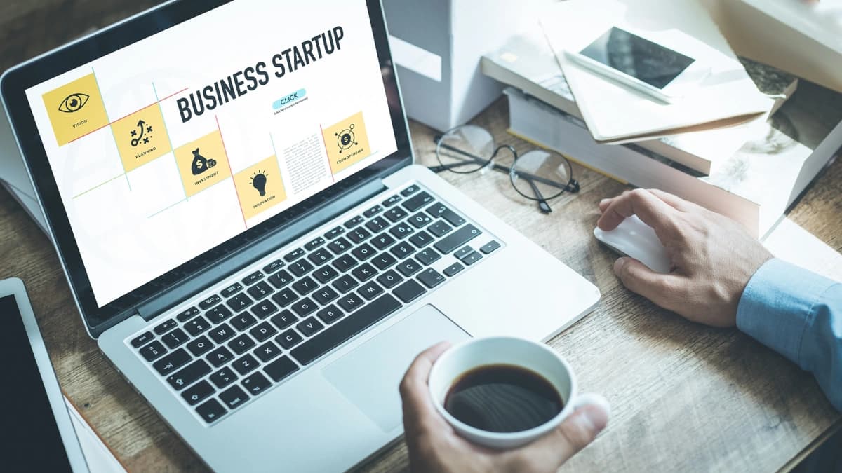 Two hands, one holding coffee and the other clicking a mouse in front of a laptop with the words "Business Startup" on the screen.