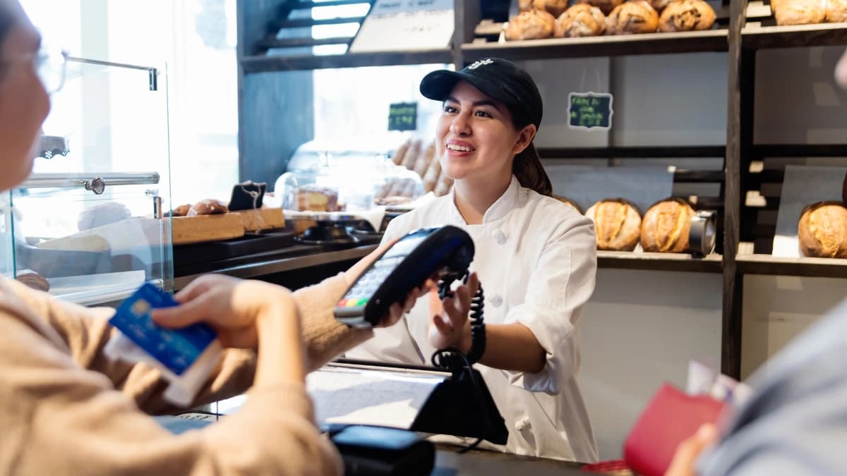 A woman working at a bakery holds out a credit card machine to a customer holding their credit card.