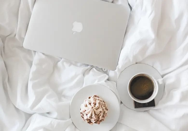 A laptop, a cup of coffee and some cream on a bed