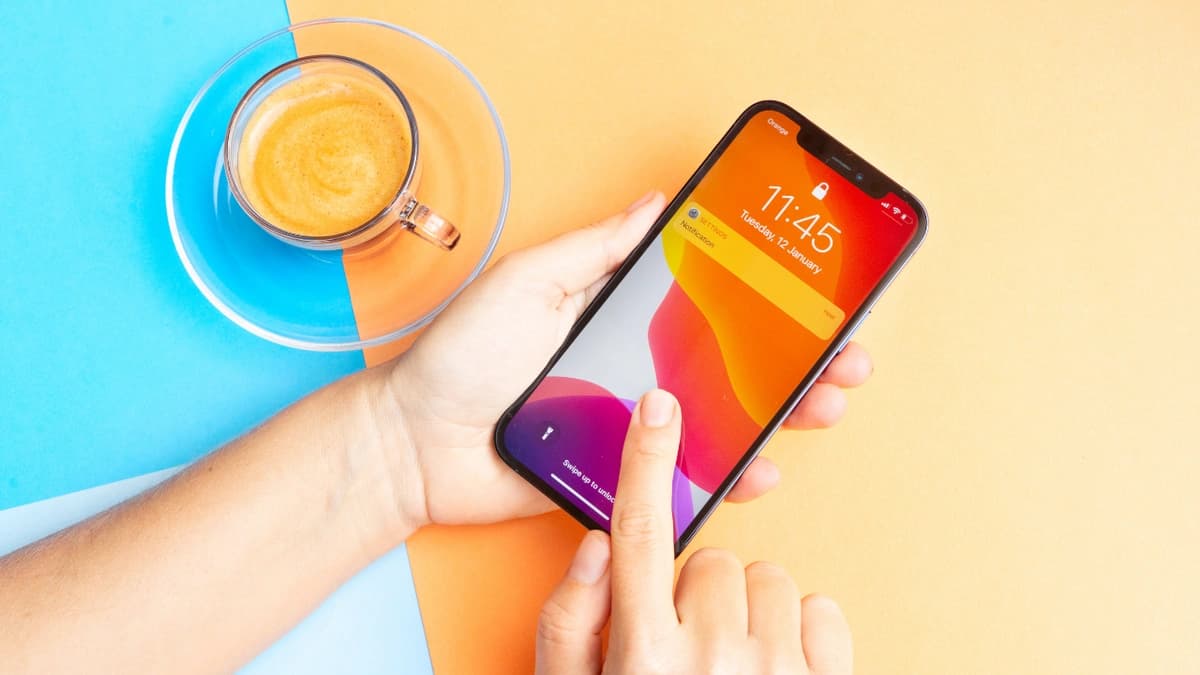 A person holding and touching their iphone, with a bright blue and orange background and a cup of coffee by their hand.