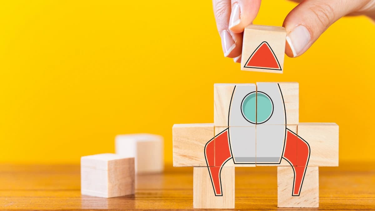 Yellow background with building blocks, a hand is stacking blocks that have the image of a rocket on them.