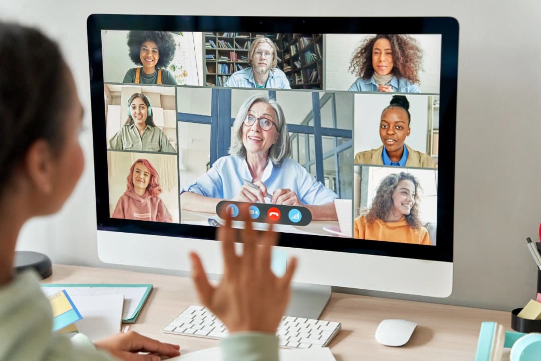 Woman looking at computer screen and waiving to a remote team of co-workers in a video call.