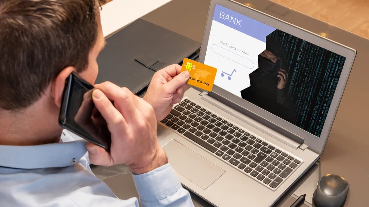 Man sitting in front of laptop while holding his credit card and a phone, giving card details to a cybercriminal who is shown on the laptop screen.